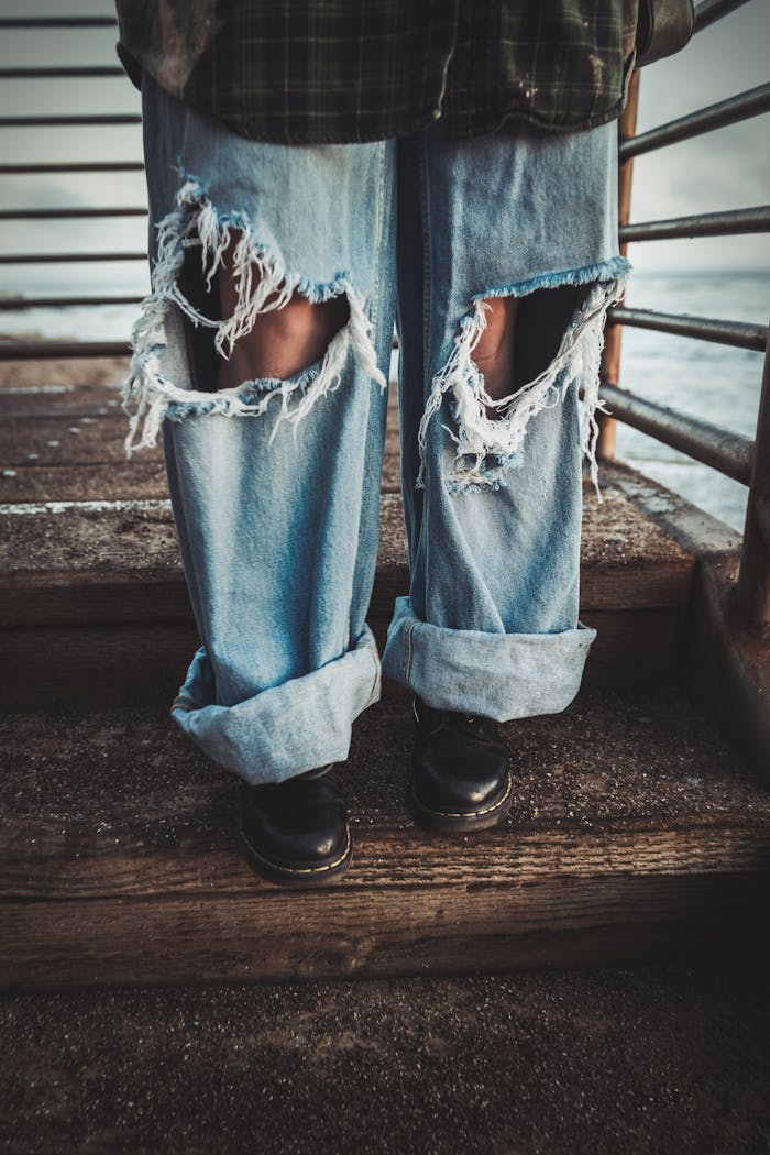 Close-up of ripped jeans and black boots on wooden stairs with metal railings.